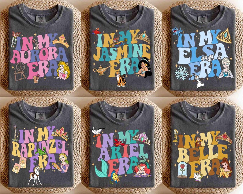 Op de afbeelding: Zes donkergrijze t-shirts met kleurrijke retro-ontwerpen. Elk shirt heeft de zin "In My" gevolgd door de naam van een prinses en "Era". De ontwerpen bevatten afbeeldingen van Aurora, Jasmine, Elsa, Rapunzel, Ariel en Belle, met bijbehorende iconen.
