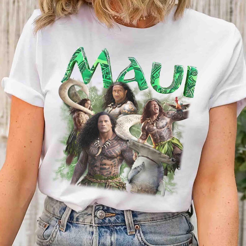 May include: A white short-sleeved t-shirt featuring a graphic print with the word Maui in large green patterned letters. The design includes multiple depictions of a muscular character with tattoos, a large fish hook, and green lightning effects.