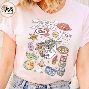 May include: Light pink t-shirt featuring a colorful graphic design with characters and elements from the animated film "Toy Story." The design includes Woody, Buzz Lightyear, and other iconic imagery. The text "Don't you get it? Friend in me" is also visible.