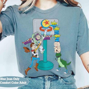 May include: Blue jean colored t-shirt featuring a basketball scene with cartoon characters Buzz Lightyear, Woody, and other Toy Story characters. The shirt has the text "Blue Jean Only With Comfort Color Adult" at the bottom.