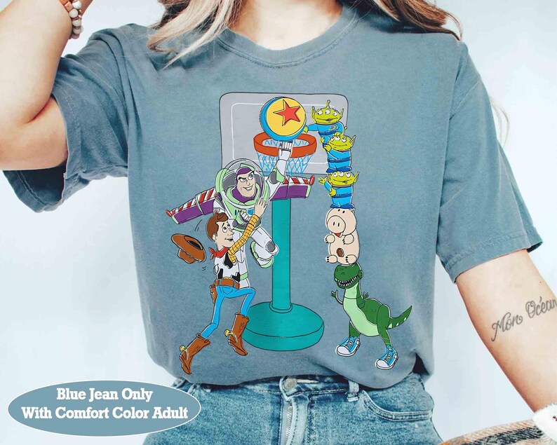 May include: Blue jean colored t-shirt featuring a basketball scene with cartoon characters Buzz Lightyear, Woody, and other Toy Story characters. The shirt has the text "Blue Jean Only With Comfort Color Adult" at the bottom.