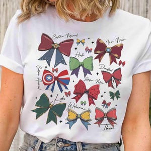 May include: White t-shirt featuring colorful bow illustrations inspired by Marvel characters, including Captain Marvel, Iron Man, and Thor. Each bow is uniquely designed with character-specific colors and details, with character names written above the bows.