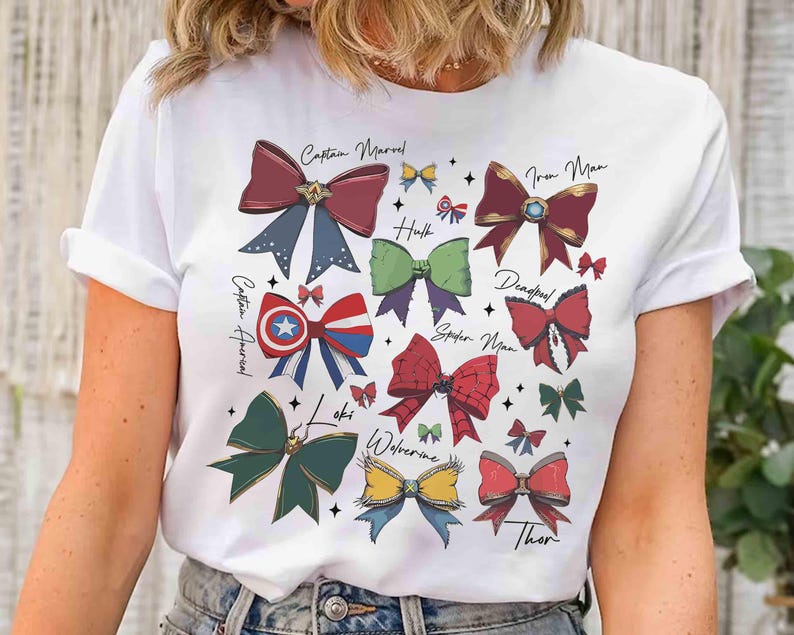 May include: White t-shirt featuring colorful bow illustrations inspired by Marvel characters, including Captain Marvel, Iron Man, and Thor. Each bow is uniquely designed with character-specific colors and details, with character names written above the bows.