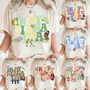 Op de afbeelding: Cr&egrave;mekleurige t-shirts met verschillende grafische ontwerpen. E&eacute;n shirt zegt "In My Tiana Era" met cartoonafbeeldingen van een kikker, een prinses en een waterlelie. Andere shirts zeggen "In My Snow White Era", "In My Cinderella Era", "In My Merida Era", "In My Moana Era" en "In My Mulan Era".
