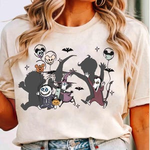 Puede incluir: Camiseta color crema con un gr&aacute;fico tem&aacute;tico de Halloween. El dise&ntilde;o incluye personajes de dibujos animados, murci&eacute;lagos, globos con caras de calaveras y Mickey Mouse, y estrellas. Los personajes est&aacute;n en varias poses, creando un aspecto l&uacute;dico y festivo.
