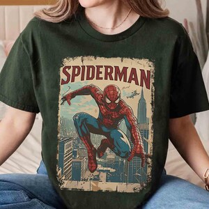 May include: Dark green t-shirt featuring a vintage-style Spider-Man graphic. The design includes the word "SPIDERMAN" in red and a detailed illustration of the superhero in action against a cityscape backdrop. The shirt is a comfortable, casual wear.