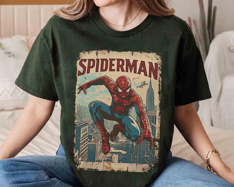 May include: Dark green t-shirt featuring a vintage-style Spider-Man graphic. The design includes the word "SPIDERMAN" in red and a detailed illustration of the superhero in action against a cityscape backdrop. The shirt is a comfortable, casual wear.
