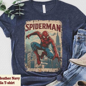 May include: Heather navy blue t-shirt featuring a vintage-style Spider-Man graphic. The design shows Spider-Man in a dynamic pose against a cityscape backdrop, with the word "SPIDERMAN" at the top. The shirt is paired with blue jeans and a pair of boots.