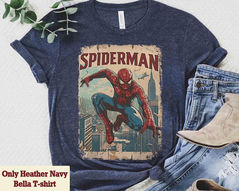 May include: Heather navy blue t-shirt featuring a vintage-style Spider-Man graphic. The design shows Spider-Man in a dynamic pose against a cityscape backdrop, with the word "SPIDERMAN" at the top. The shirt is paired with blue jeans and a pair of boots.
