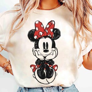May include: Cream-colored t-shirt featuring a black and white Minnie Mouse graphic. Minnie Mouse is depicted with a red polka-dot bow, black ears, and a smiling face. The design is a classic cartoon style.
