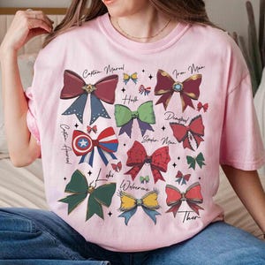 May include: Pink t-shirt featuring colorful bow designs representing Marvel characters like Captain Marvel, Iron Man, and Thor. Each bow has a unique color scheme and is labeled with the character's name. The shirt is a casual wear item.