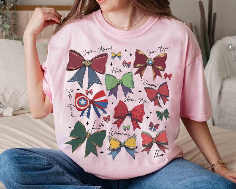 May include: Pink t-shirt featuring colorful bow designs representing Marvel characters like Captain Marvel, Iron Man, and Thor. Each bow has a unique color scheme and is labeled with the character's name. The shirt is a casual wear item.