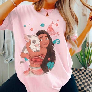 Puede incluir: Una camiseta rosa claro con una ilustraci&oacute;n gr&aacute;fica de un personaje de pelo largo abrazando a un cerdito. El dise&ntilde;o incluye elementos decorativos como flores tropicales, hojas y una forma de vela estampada en tonos rojos y azul turquesa.