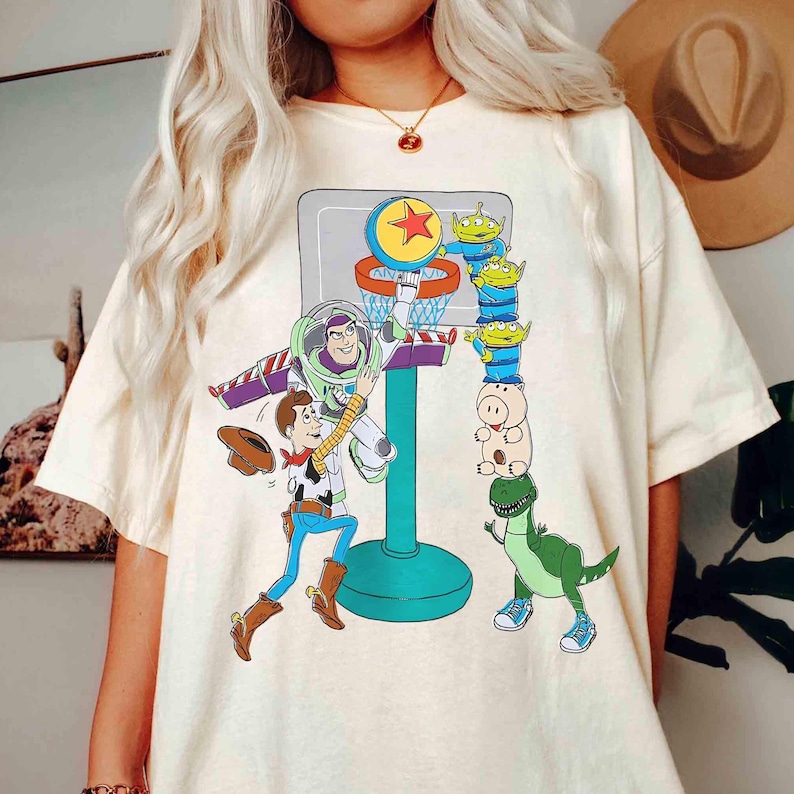 May include: Cream-colored t-shirt featuring a colorful graphic of Toy Story characters playing basketball. Buzz Lightyear is dunking the ball, while Woody and other characters watch. The design includes a basketball hoop and a star.