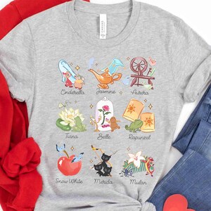 May include: A light gray t-shirt featuring colorful illustrations of Disney princesses and their associated icons, including Cinderella, Jasmine, Aurora, Tiana, Belle, Rapunzel, Snow White, Merida, and Mulan. The names of the princesses are printed below each image.