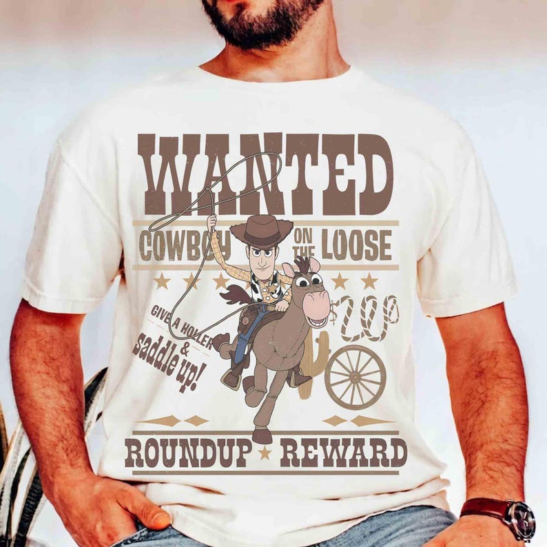 Pode incluir: Camiseta creme com um desenho de cowboy de desenho animado montado a cavalo, com o texto castanho "WANTED COWBOY ON THE LOOSE". O design inclui um la&ccedil;o, estrelas e as palavras "Give a holler & saddle up!" e "Roundup Reward".