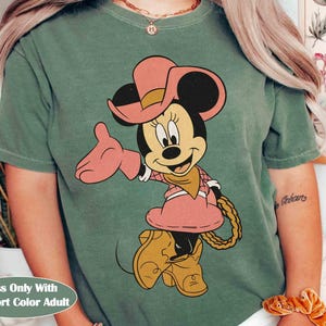 Puede incluir: Camiseta verde musgo con Minnie Mouse de dibujos animados con un atuendo de vaquera rosa, que incluye sombrero, vestido y botas. El texto "Moss Only With Comfort Color Adult" est&aacute; en la parte inferior. El dise&ntilde;o est&aacute; centrado en la camiseta.
