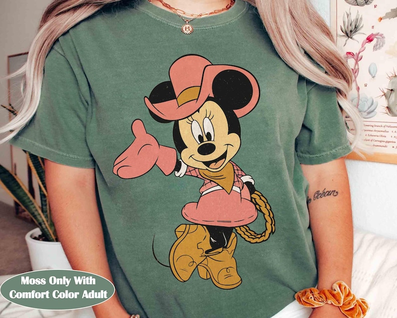 Puede incluir: Camiseta verde musgo con Minnie Mouse de dibujos animados con un atuendo de vaquera rosa, que incluye sombrero, vestido y botas. El texto "Moss Only With Comfort Color Adult" est&aacute; en la parte inferior. El dise&ntilde;o est&aacute; centrado en la camiseta.