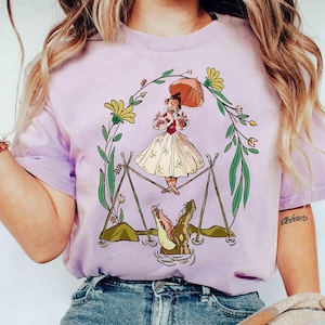 Puede incluir: Una camiseta lila con una ilustración de dibujos animados de una mujer con un paraguas, de pie sobre caimanes. El diseño está enmarcado por una corona floral con flores amarillas y hojas verdes. La mujer lleva un vestido blanco y sostiene un paraguas rosa.