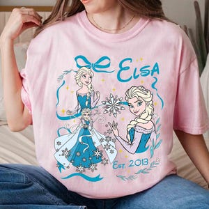 K&ouml;nnte beinhalten: Hellrosa T-Shirt mit einem Elsa-Motiv aus Frozen. Das Design zeigt Illustrationen von Elsa in einem blauen Kleid, Schneeflocken und den Text "Elsa Est. 2013" in Blau. Das T-Shirt hat einen Rundhalsausschnitt.