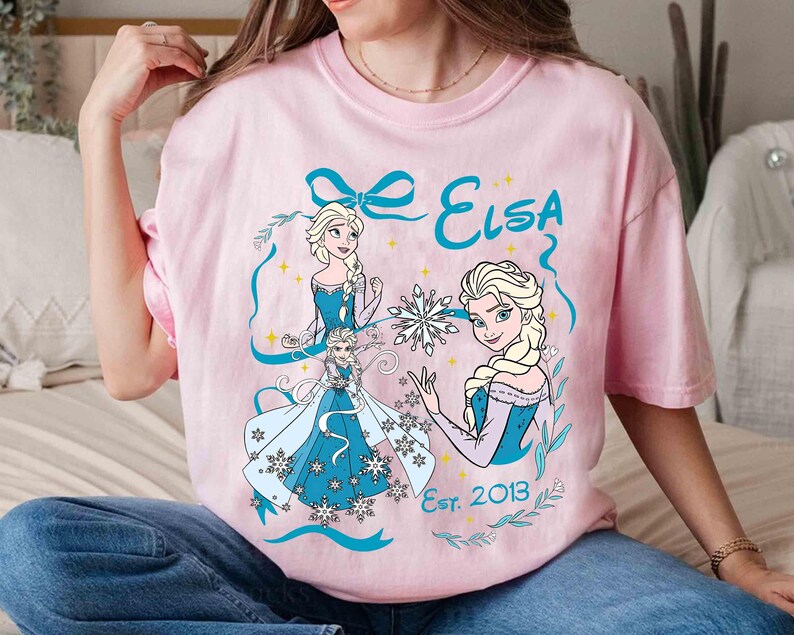 K&ouml;nnte beinhalten: Hellrosa T-Shirt mit einem Elsa-Motiv aus Frozen. Das Design zeigt Illustrationen von Elsa in einem blauen Kleid, Schneeflocken und den Text "Elsa Est. 2013" in Blau. Das T-Shirt hat einen Rundhalsausschnitt.