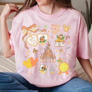 May include: Pink t-shirt featuring a colorful graphic design with the text "Sunshine On my mind". The design includes an orange character, a castle, fireworks, and other whimsical elements. The shirt is a light pink color.