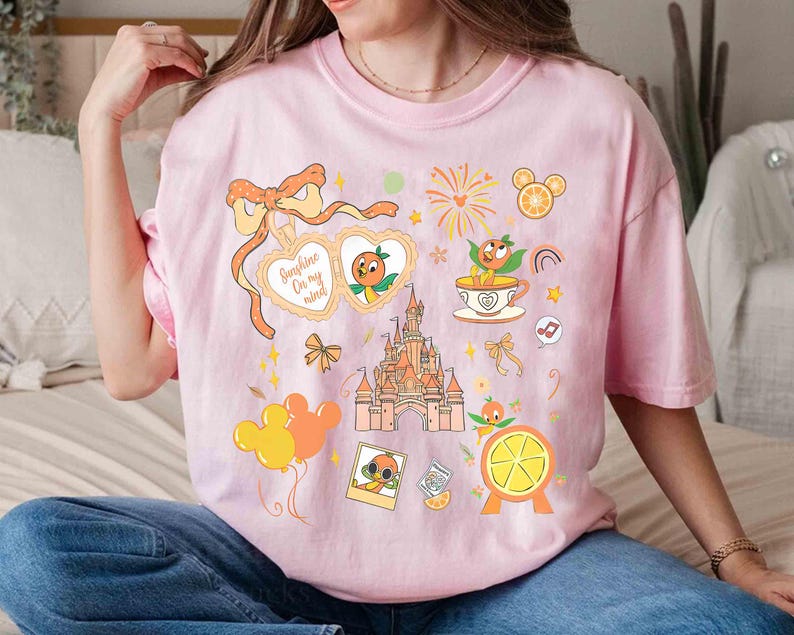 May include: Pink t-shirt featuring a colorful graphic design with the text "Sunshine On my mind". The design includes an orange character, a castle, fireworks, and other whimsical elements. The shirt is a light pink color.