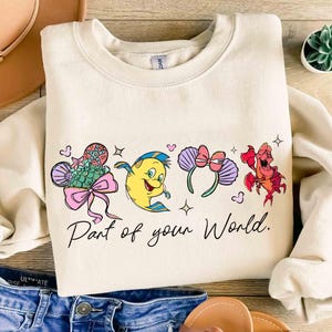 Puede incluir: Sudadera crema con personajes de dibujos animados coloridos y el texto "Part of your World". El dise&ntilde;o incluye un pez, un personaje amarillo, una diadema con lazo y un cangrejo rojo. La sudadera se combina con pantalones cortos de mezclilla y un sombrero.