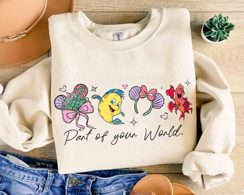 Puede incluir: Sudadera crema con personajes de dibujos animados coloridos y el texto "Part of your World". El dise&ntilde;o incluye un pez, un personaje amarillo, una diadema con lazo y un cangrejo rojo. La sudadera se combina con pantalones cortos de mezclilla y un sombrero.
