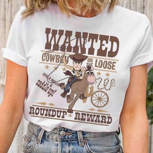 Pode incluir: Camiseta branca com um cowboy de desenho animado montado a cavalo e o texto castanho "WANTED COWBOY ON THE LOOSE". O design inclui estrelas, uma roda de carro e as palavras "ROUNDUP REWARD".
