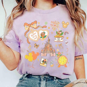 May include: A light purple t-shirt featuring a collection of orange-themed illustrations. The design includes a castle, orange slices, a character in sunglasses, and the text "Sunshine On my mind". The shirt has a relaxed fit.