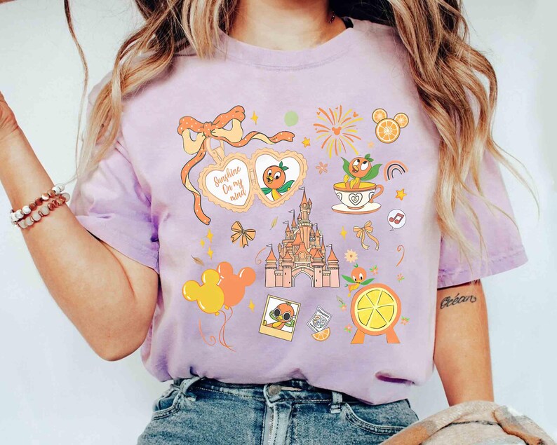 May include: A light purple t-shirt featuring a collection of orange-themed illustrations. The design includes a castle, orange slices, a character in sunglasses, and the text "Sunshine On my mind". The shirt has a relaxed fit.