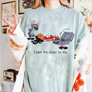 May include: A light blue t-shirt featuring a cartoon illustration of Lilo and Stitch. Stitch stands near Lilo, who is lying down next to a record player. The text "Leave me alone to die" is printed on the shirt.