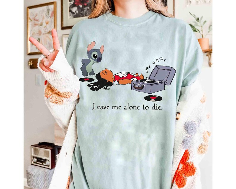 May include: A light blue t-shirt featuring a cartoon illustration of Lilo and Stitch. Stitch stands near Lilo, who is lying down next to a record player. The text "Leave me alone to die" is printed on the shirt.