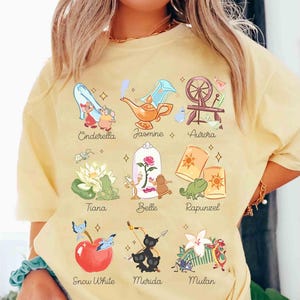 May include: A light yellow t-shirt featuring illustrations of Disney princesses, including Cinderella, Jasmine, Aurora, Tiana, Belle, Rapunzel, Snow White, Merida, and Mulan. Each princess is depicted with a related icon and name.