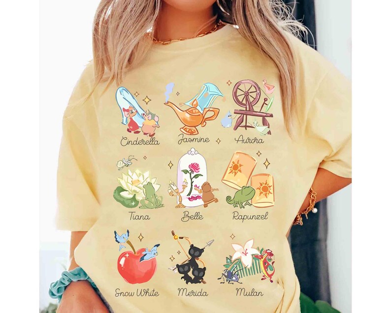 May include: A light yellow t-shirt featuring illustrations of Disney princesses, including Cinderella, Jasmine, Aurora, Tiana, Belle, Rapunzel, Snow White, Merida, and Mulan. Each princess is depicted with a related icon and name.