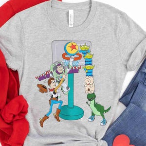 May include: A light gray t-shirt featuring a colorful graphic of Toy Story characters playing basketball. Buzz Lightyear is dunking the ball, while Woody and other characters watch. The shirt is made of a soft material.