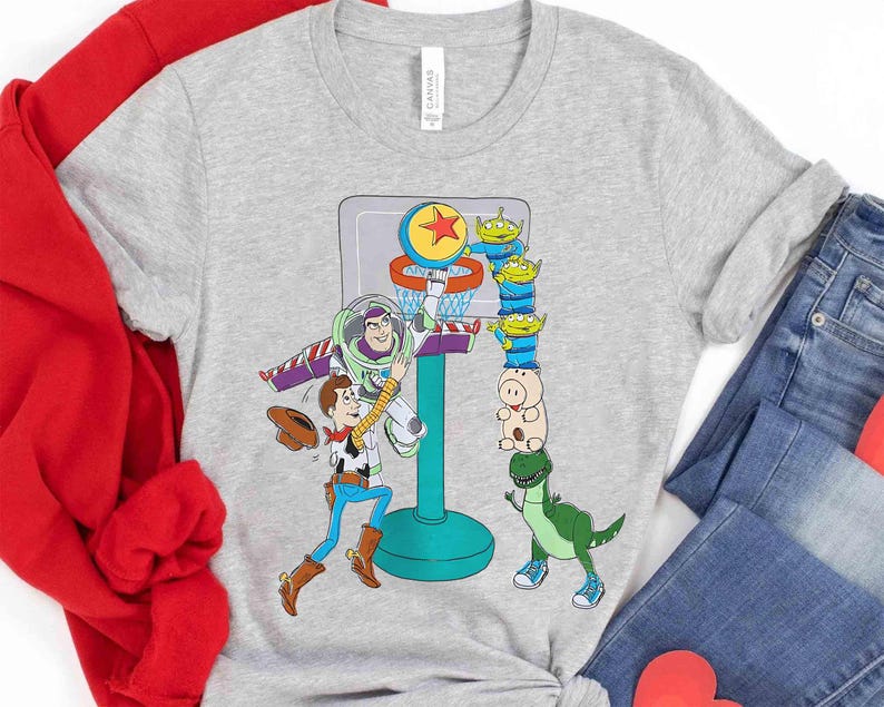 May include: A light gray t-shirt featuring a colorful graphic of Toy Story characters playing basketball. Buzz Lightyear is dunking the ball, while Woody and other characters watch. The shirt is made of a soft material.