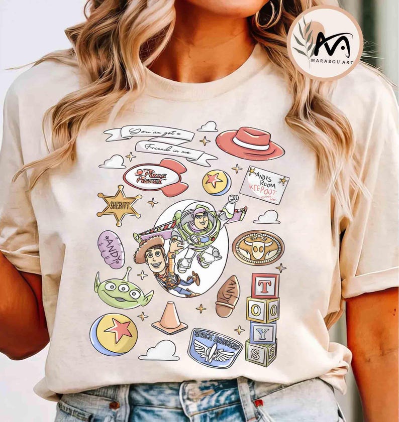 May include: Beige t-shirt featuring a colorful graphic of Toy Story characters and elements. The design includes Buzz Lightyear, Woody, and various themed icons like a sheriff badge, a hat, and the text "You've got a friend in me."