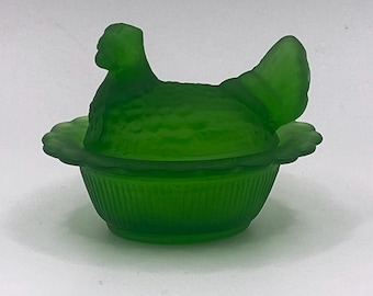 May include: A green glass hen-shaped covered dish. The hen sits atop a scalloped-edged bowl with vertical ribbed detailing. The hen's body is textured, and the lid forms the hen's head and wings. A decorative kitchenware item.