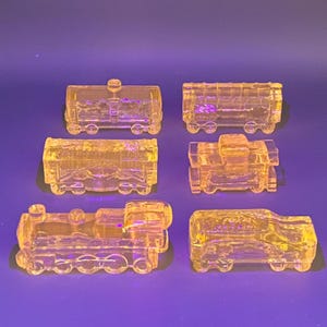 May include: Six yellow glass train car figurines are arranged on a purple surface. The set includes a locomotive, a caboose, and four different types of freight cars. Each piece has detailed molding and sits on small wheels.