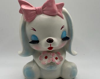 May include: A ceramic bunny figurine with a large pink bow on its head. The bunny has blue eyes, black eyelashes, and a red mouth. It holds a bouquet of pink flowers. The bunny is white with blue accents.