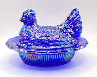 May include: A blue iridescent hen-shaped covered dish. The hen sits atop a scalloped-edge dish. The dish and hen have a textured surface and a vibrant, color-shifting finish. A decorative kitchenware item.