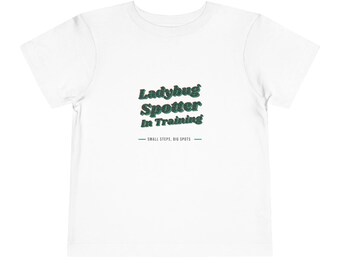 May include: A white short-sleeved t-shirt featuring the text Ladybug Spotter In Training in a dark green font with a subtle red outline. Below the main text, the phrase SMALL STEPS, BIG SPOTS is printed in a smaller, dark green sans-serif font.