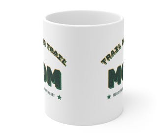 May include: A white ceramic mug featuring the text TRAIL DOG MOM in dark green block letters with a textured appearance. The design includes the words MUDDY PAWS HAPPY HEART and a small green star, creating a clear and legible graphic on the mug.
