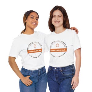 Pu&ograve; includere: Una t-shirt bianca a maniche corte con una grafica circolare che include una piccola icona a forma di uovo, il testo "JUST ONE MORE HEN, I PROMISE" lungo il bordo superiore e un banner arancione al centro con il testo "Chicken Math Is Real".