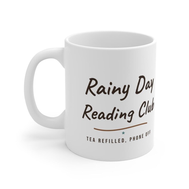 May include: A white ceramic mug with a handle featuring the dark brown text Rainy Day Reading Club in a script font, followed by a horizontal line, a small teal star, and the text TEA REFILLED, PHONE OFF in a simple sans-serif font.