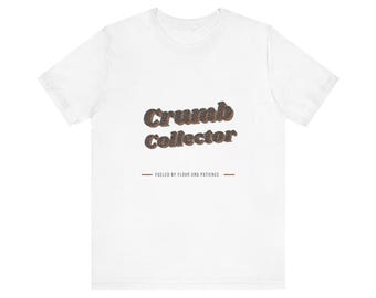 Può includere: Una t-shirt bianca a maniche corte con la scritta Crumb Collector in un carattere scuro e testurizzato. Sotto il testo principale, una riga più piccola riporta FUELED BY FLOOR AND PATIENCE, affiancata da due linee orizzontali sul petto.