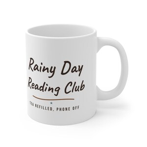 May include: A white ceramic mug featuring the dark brown text Rainy Day Reading Club in a handwritten script font, followed by a horizontal line, a small teal star, and the text TEA REFILLED, PHONE OFF in a simple sans-serif font.