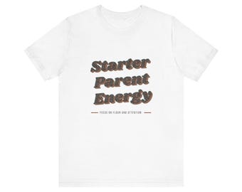 Può includere: Una t-shirt bianca a maniche corte con il testo Starter Parent Energy in un carattere scuro, testurizzato e maculato. Sotto il testo principale, una riga più piccola riporta FEEDS ON FLOUR AND ATTENTION, fiancheggiata da due linee orizzontali.